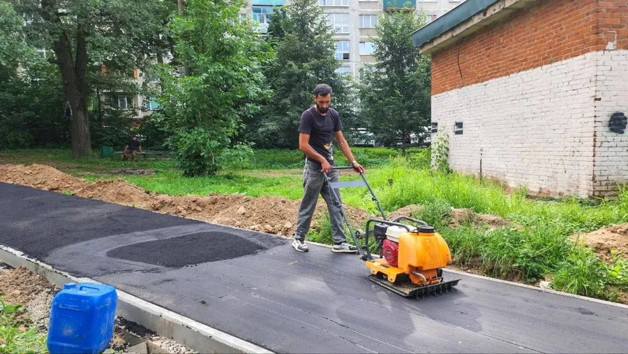 Рабочий уплотняет виброплитой свежеуложенный асфальт пешеходной дорожки